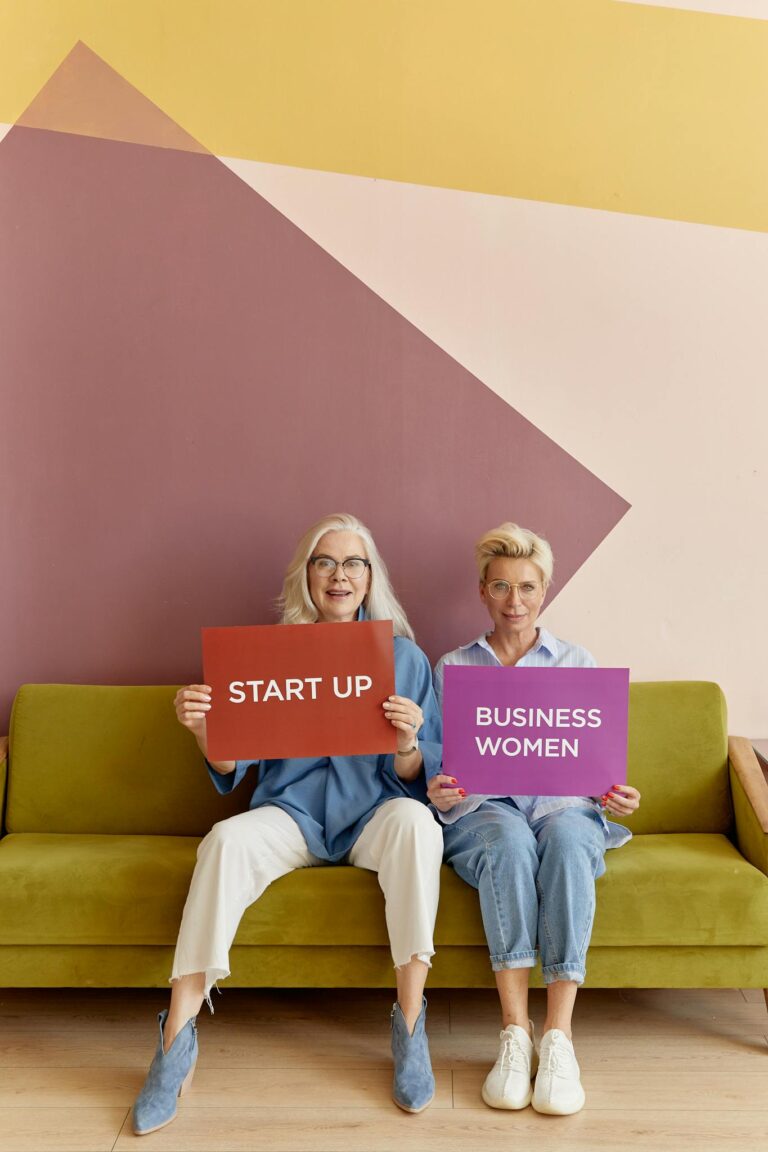 Two senior women holding startup and businesswomen signs, sitting on a sofa.