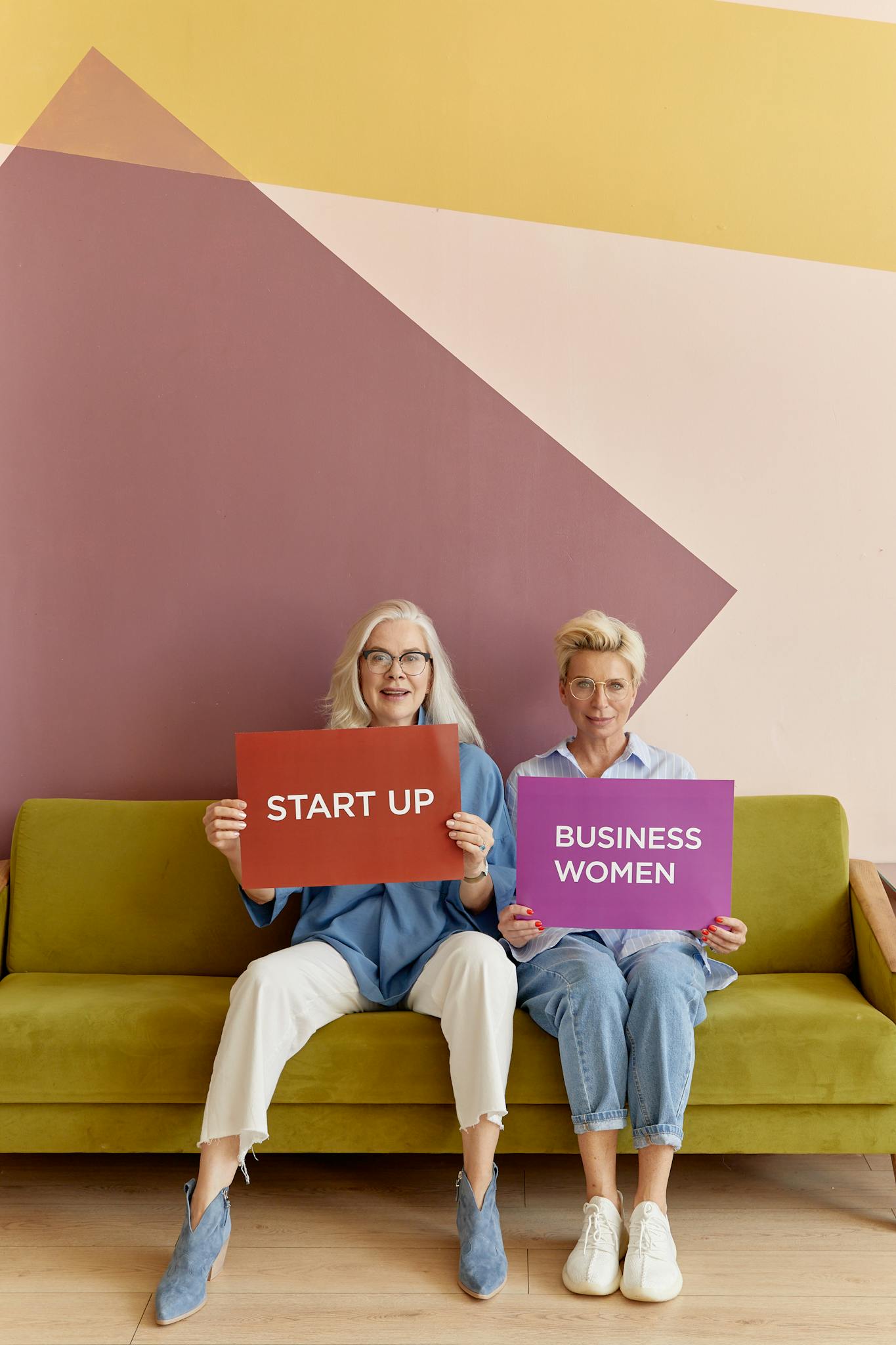 Two senior women holding startup and businesswomen signs, sitting on a sofa.